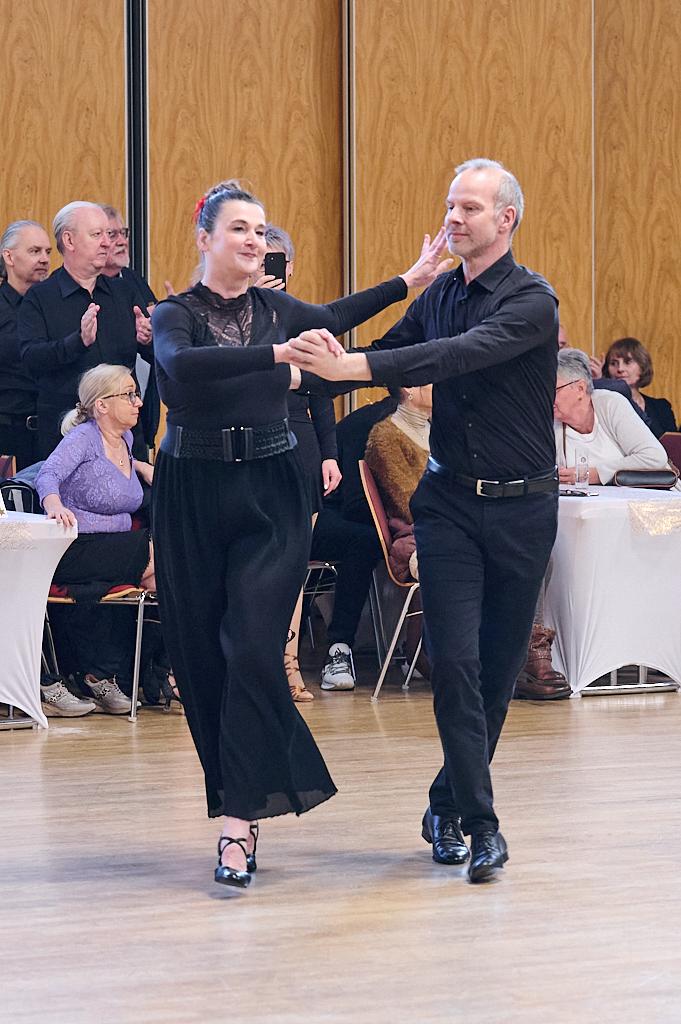 Tanzpaar in eleganter Kleidung beim Tanzen auf einer Veranstaltung, Zuschauer beobachten im Hintergrund.