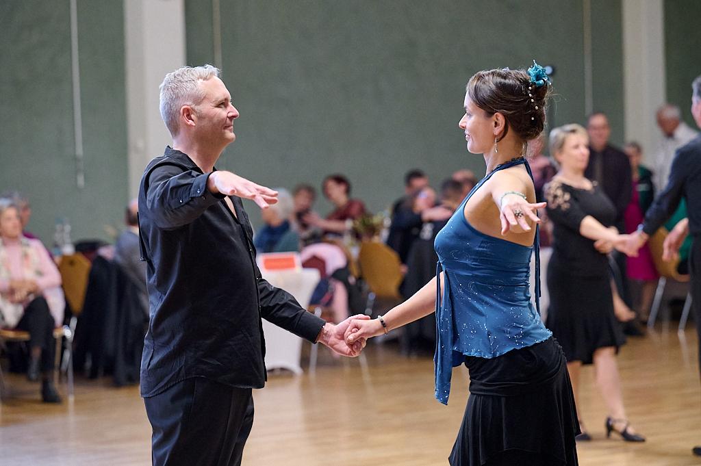 Tanzpaar in eleganter Kleidung beim Gesellschaftstanz in einem festlichen Saal mit anderen T&auml;nzern im Hintergrund.