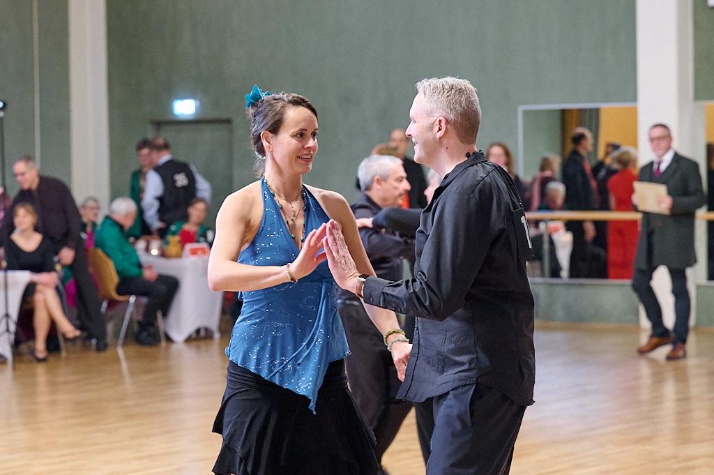 T&auml;nzerpaar in festlicher Kleidung beim Gesellschaftstanz in einem Saal mit mehreren Zuschauern im Hintergrund.