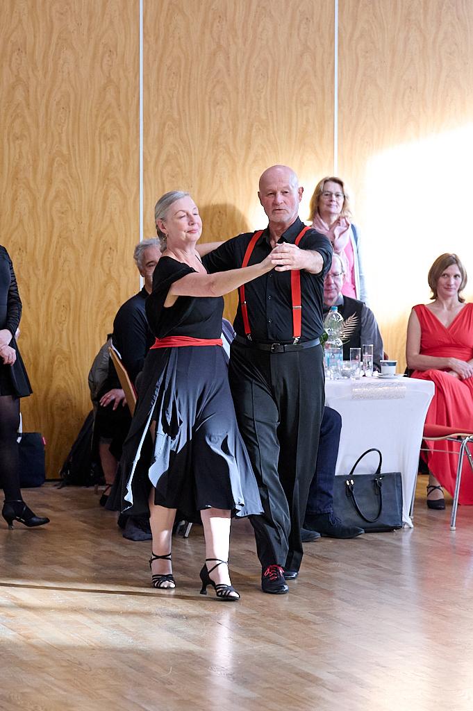 Tanzendes Paar in eleganter Kleidung, umgeben von Zuschauern in festlicher Atmosph&auml;re. Holzfu&szlig;boden im Hintergrund.