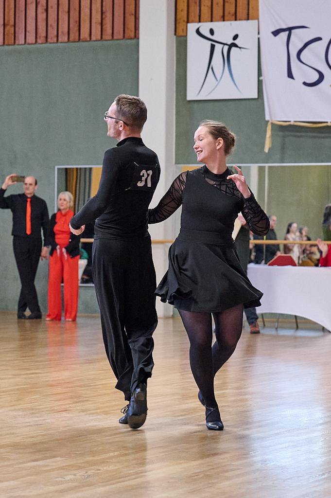 Tanzpaar in eleganter Kleidung w&auml;hrend eines Wettbewerbs auf einem Parkettboden, umgeben von Zuschauern.