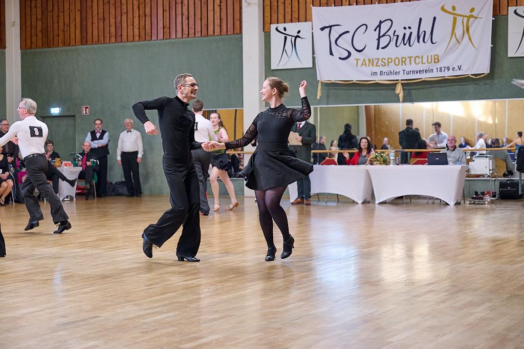 Tanzpaar in eleganter Kleidung f&uuml;hrt einen Kontakt-Tanz in einem Saal mit Zuschauern im Hintergrund aus.