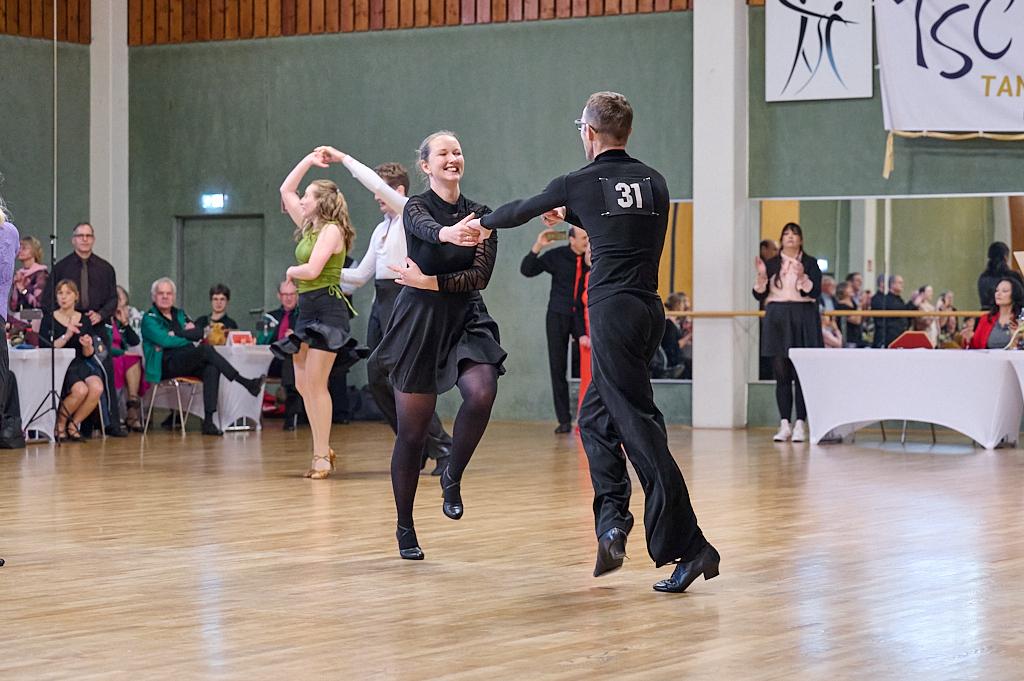 Tanzpaare in eleganter Kleidung beim Wettkampf auf einer Tanzfl&auml;che, Zuschauer im Hintergrund applaudieren.