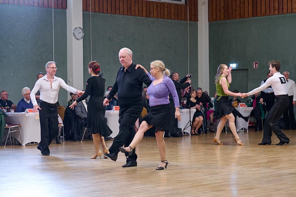 Tanzpaar beim Gesellschaftstanz in formeller Kleidung auf einer hellen Tanzfl&auml;che, im Hintergrund Zuschauer und Tische.