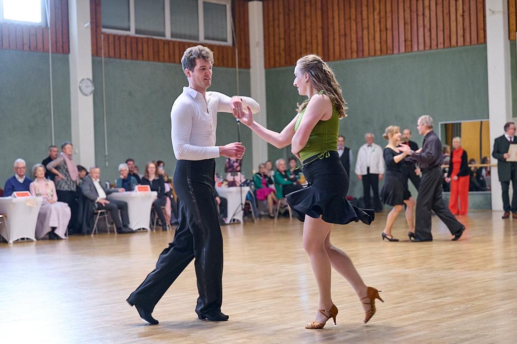 Tanzpaar in eleganter Kleidung, das in einem Saal zu Musik tanzt, Zuschauer im Hintergrund.
