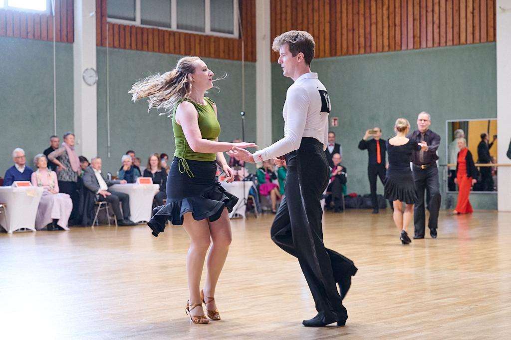 Tanzendes Paar in fr&ouml;hlicher Pose auf einer Tanzfl&auml;che, Zuschauer im Hintergrund beobachten die Darbietung.
