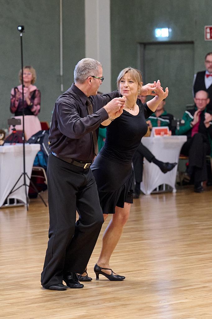Tanzpaar in eleganter Kleidung tanzt auf einer Holzfl&auml;che, im Hintergrund applaudierende Zuschauer.