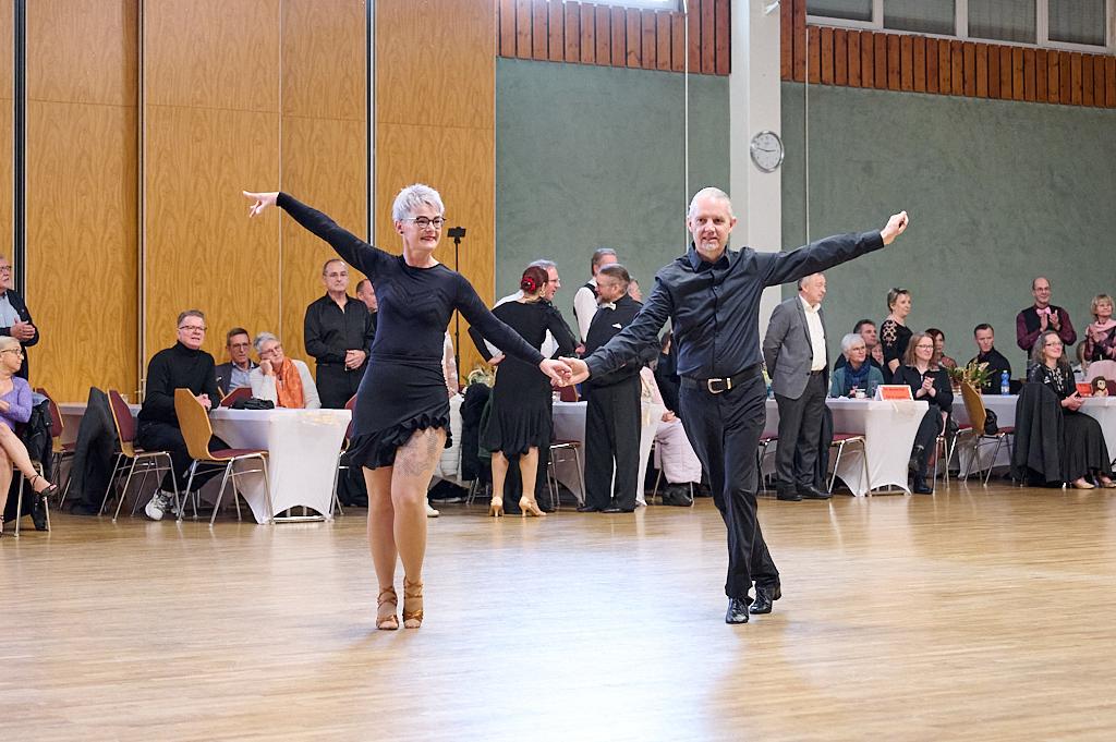 Tanzpaar in eleganter Kleidung f&uuml;hrt einen Tanz auf einer Holzparkettfl&auml;che vor einem Publikum aus mehreren Menschen vor.