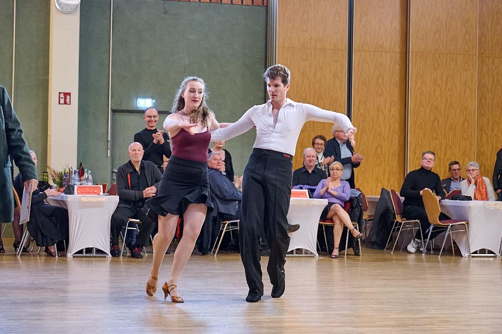 Tanzpaar in eleganter Kleidung f&uuml;hrt einen Tanz auf einer B&uuml;hnenfl&auml;che vor applaudierendem Publikum aus.