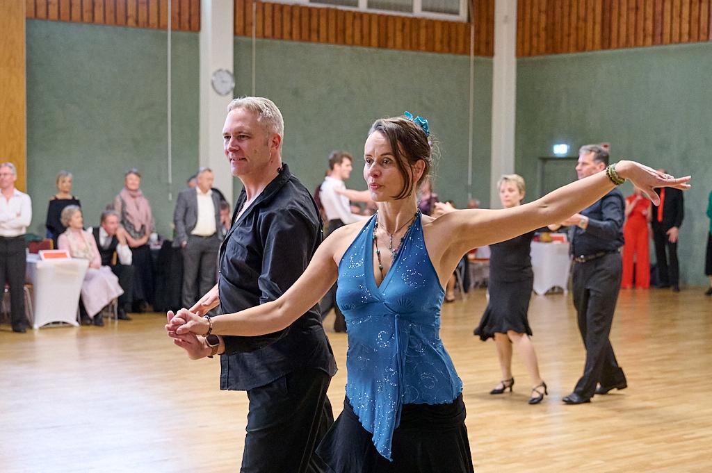 Tanzpaar f&uuml;hrt einen leidenschaftlichen Tanz in einem festlichen Saal mit tanzenden G&auml;sten im Hintergrund aus.