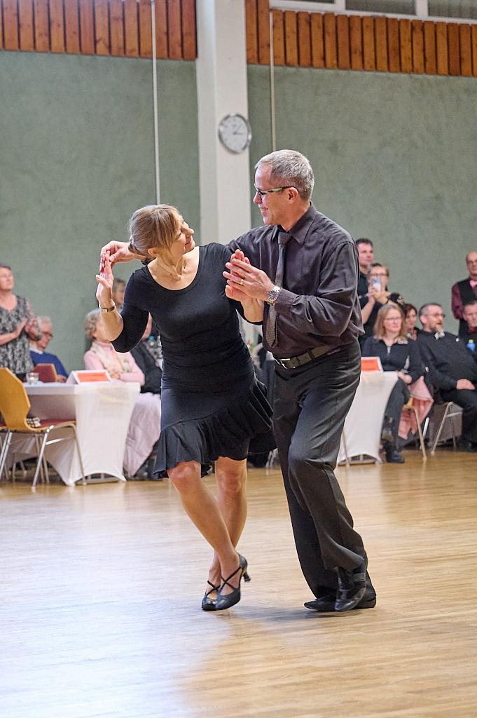 Tanzpaar in eleganter Kleidung w&auml;hrend einer Tanzveranstaltung auf einem Holzfu&szlig;boden, Zuschauer im Hintergrund.