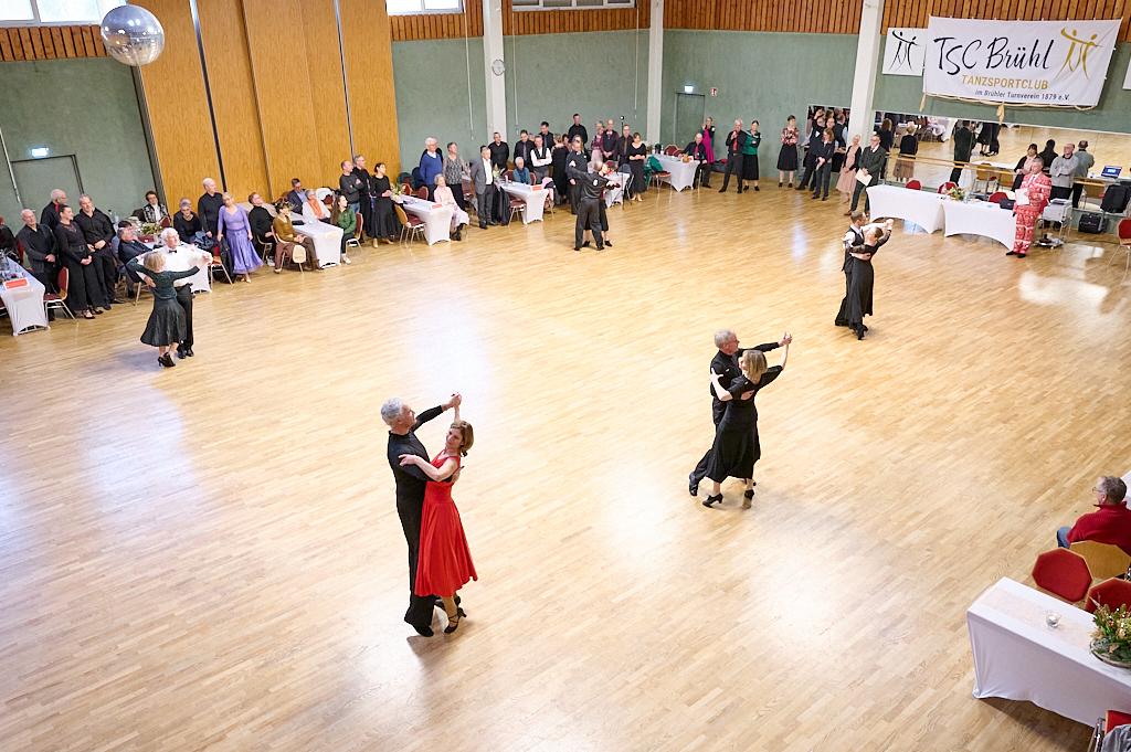Tanzende Paare in eleganter Kleidung auf einer Holzbodenfl&auml;che, umgeben von Zuschauern und festlicher Dekoration.