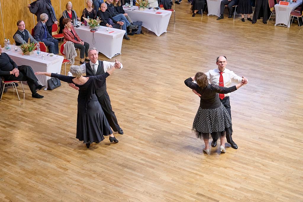 Tanzpaar mit eleganter Kleidung tanzt auf einem Parkettboden, im Hintergrund Zuschauer an Tischen.