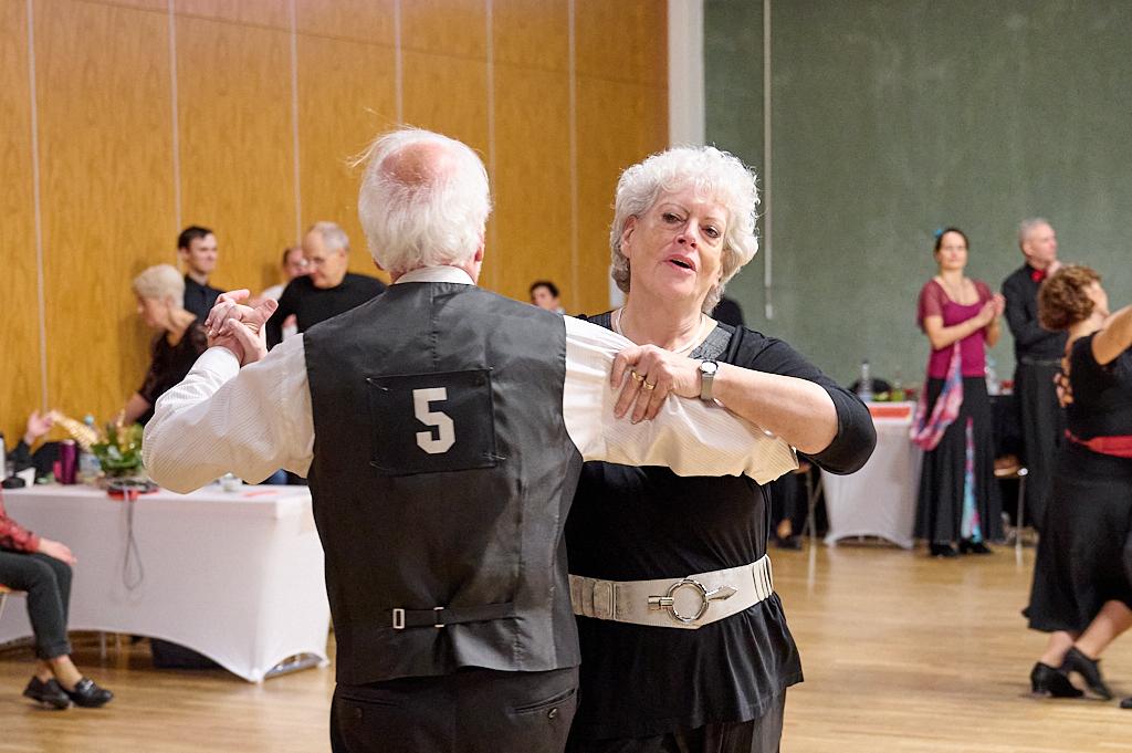 Tanzpaar in eleganter Kleidung bei einer Veranstaltung, umgeben von weiteren T&auml;nzern in einem festlichen Saal.