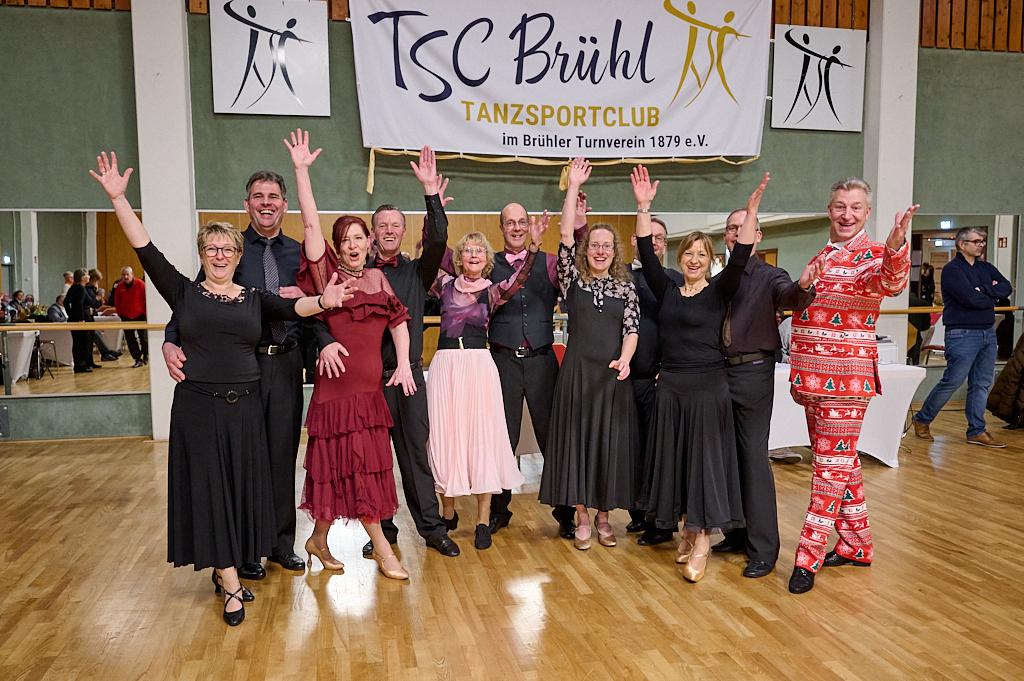 Eine Gruppe von T&auml;nzern in formeller Kleidung posiert fr&ouml;hlich vor einem Banner des TSC Br&uuml;hl in einer Tanzhalle.
