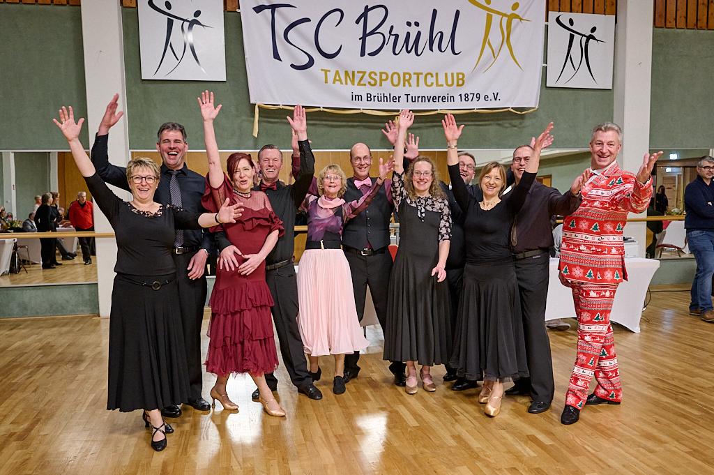 Gruppe von T&auml;nzern in festlicher Kleidung mit erhobenen H&auml;nden vor einem Banner des TSC Br&uuml;hl in einem Tanzsaal.