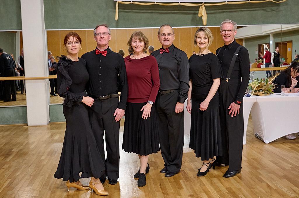 Sechs Tanzpaare stehen in eleganter Kleidung, vor einem Spiegel und einer Tanzfl&auml;che in einem hellen Saal.