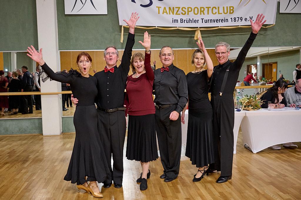 Sechs tanzende Personen in eleganter Kleidung jubeln in einem Tanzsaal mit Spiegelwand und festlicher Dekoration.