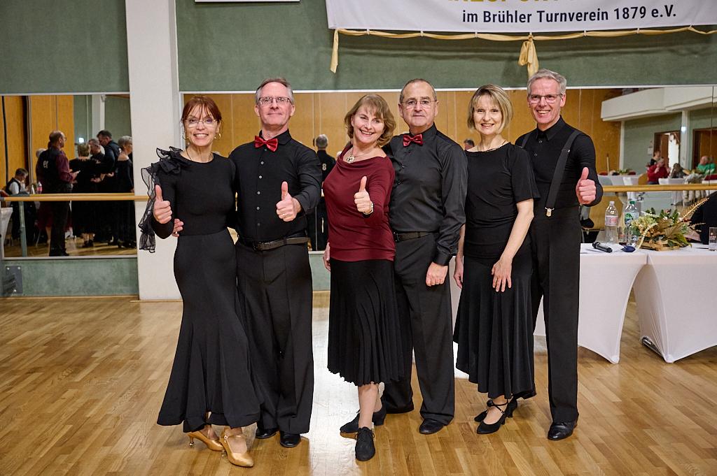 Sechs T&auml;nzer in eleganter Kleidung geben Daumen hoch auf einer Tanzveranstaltung, im Hintergrund eine Tanzfl&auml;che.