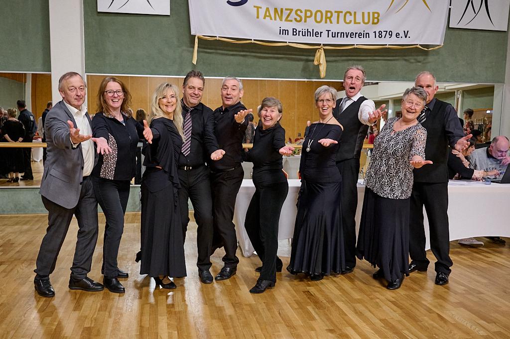Gruppe von tanzenden Menschen in schwarz, l&auml;chelnd und mit offenen H&auml;nden, in einem Tanzsaal mit Spiegelw&auml;nden.