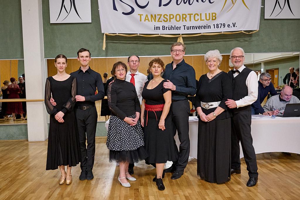 Gruppe von Tanzsportlern in formeller Kleidung steht vor einem Spiegel, im Hintergrund ein Tisch mit Computern.