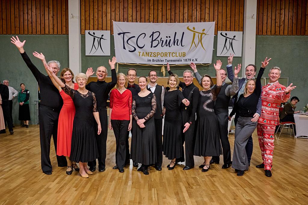 Gruppenselfie von T&auml;nzern des TSC Br&uuml;hl in festlicher Kleidung, l&auml;chelnd vor einem Vereinsbanner.