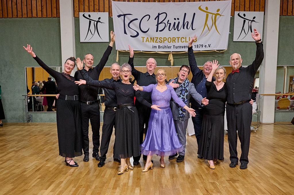 Gruppenszene mit T&auml;nzern in festlicher Kleidung, die vor einem Banner des TSC Br&uuml;hl posieren und Freude zeigen.