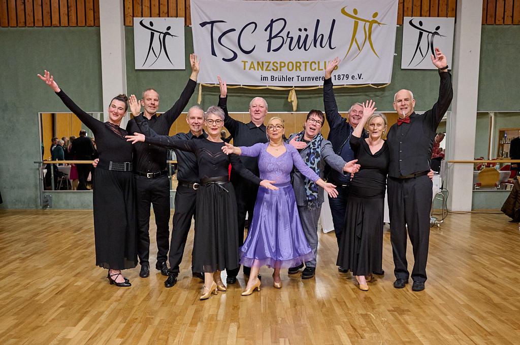Gruppe von T&auml;nzern in eleganter Abendkleidung, die freudig in einem Tanzsaal posieren, vor einem Banner des Tanzsportclubs.