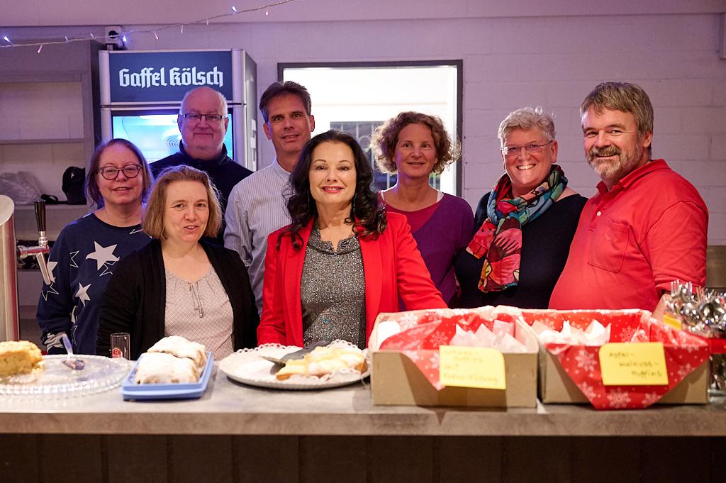 Gruppenshot von acht Personen l&auml;chelnd hinter einem Tisch mit Geb&auml;ck und Snacks in einem festlichen Raum.