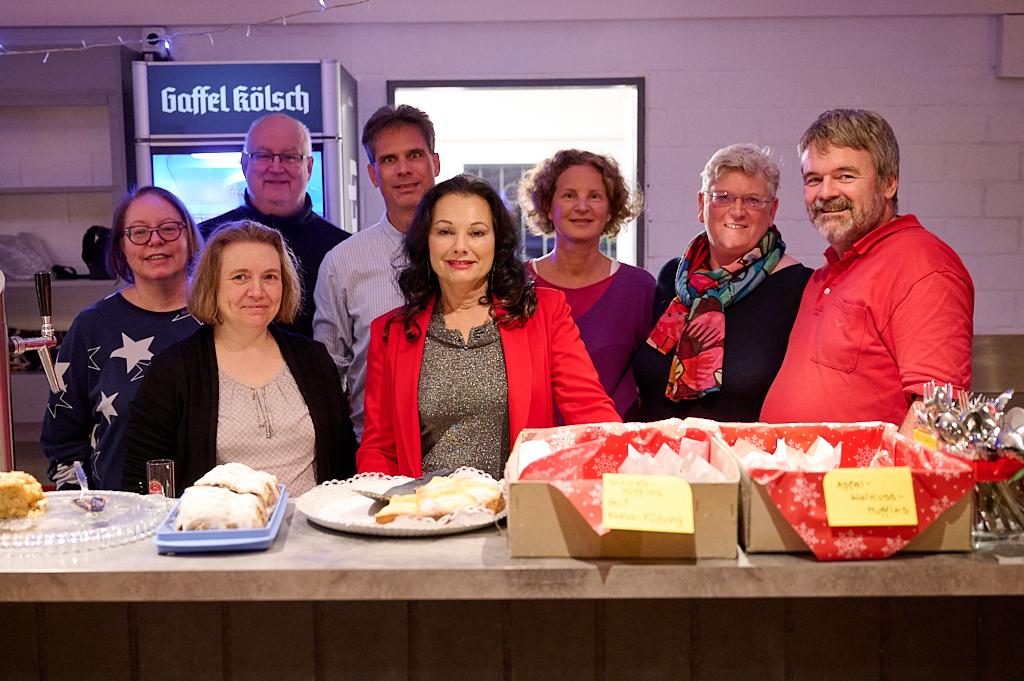 Gruppe von acht Personen l&auml;chelt vor einem Stehtisch mit verschiedenen Speisen und Getr&auml;nken.