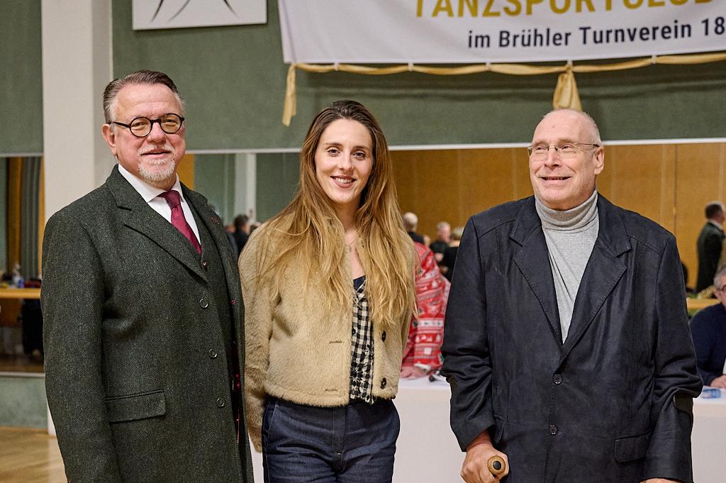 Drei Personen posieren l&auml;chelnd vor einem Banner im Hintergrund, das auf einen Tanzsportverein hinweist.