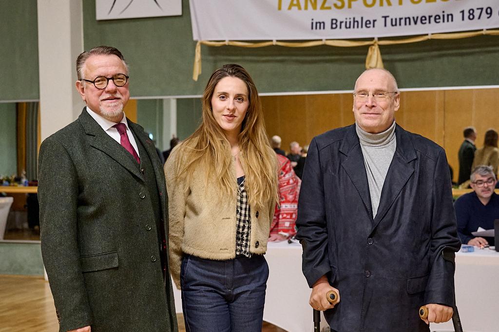 Drei Personen stehen l&auml;chelnd in einem Raum mit festlicher Dekoration im Hintergrund.