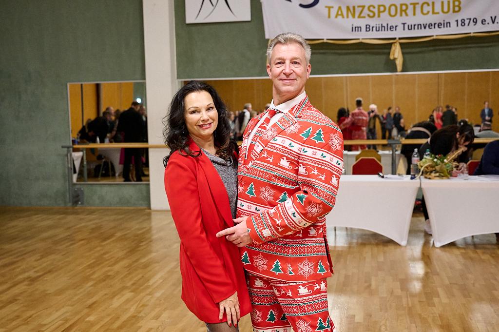 Paar l&auml;chelt in festlicher Kleidung, der Mann tr&auml;gt einen bunten Anzug mit Weihnachtsmotiven, im Hintergrund Tanzveranstaltung.