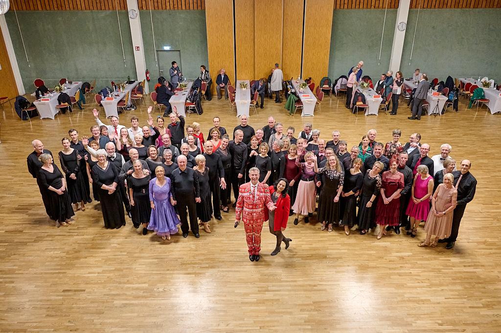 Gruppensfoto von T&auml;nzern in eleganter Kleidung, die auf einer Tanzfl&auml;che zusammenstehen. Tische im Hintergrund.