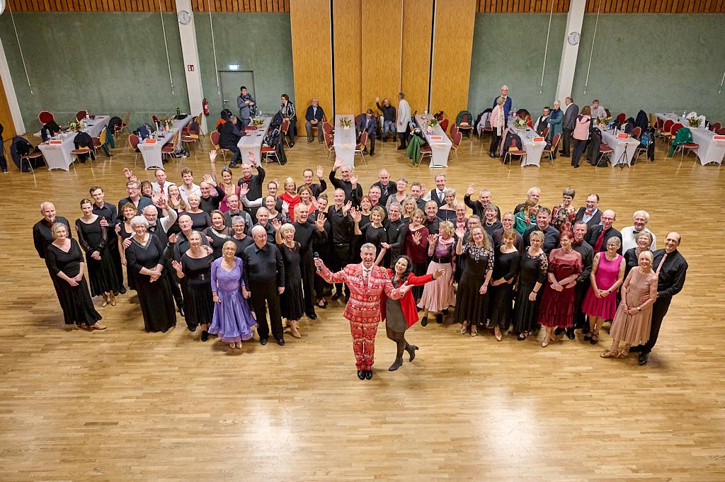 Gruppengestellte T&auml;nzer in festlicher Kleidung l&auml;cheln und winken in einer gro&szlig;en Tanzfl&auml;che mit Tischdekoration.