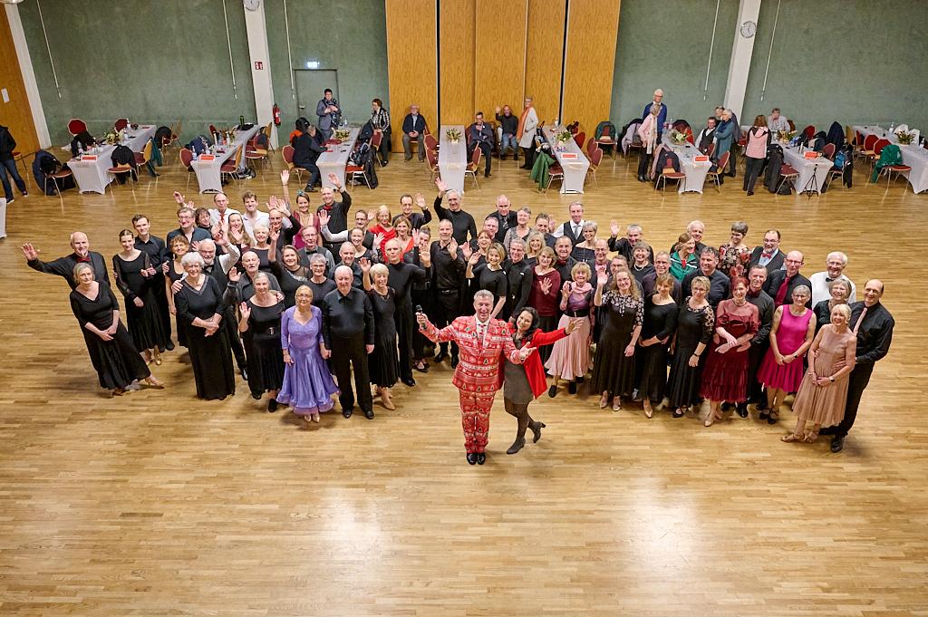 Gruppierung von etwa 60 Personen in eleganter Tanzkleidung, einige winken, in einem festlichen Saal mit Tischen im Hintergrund.