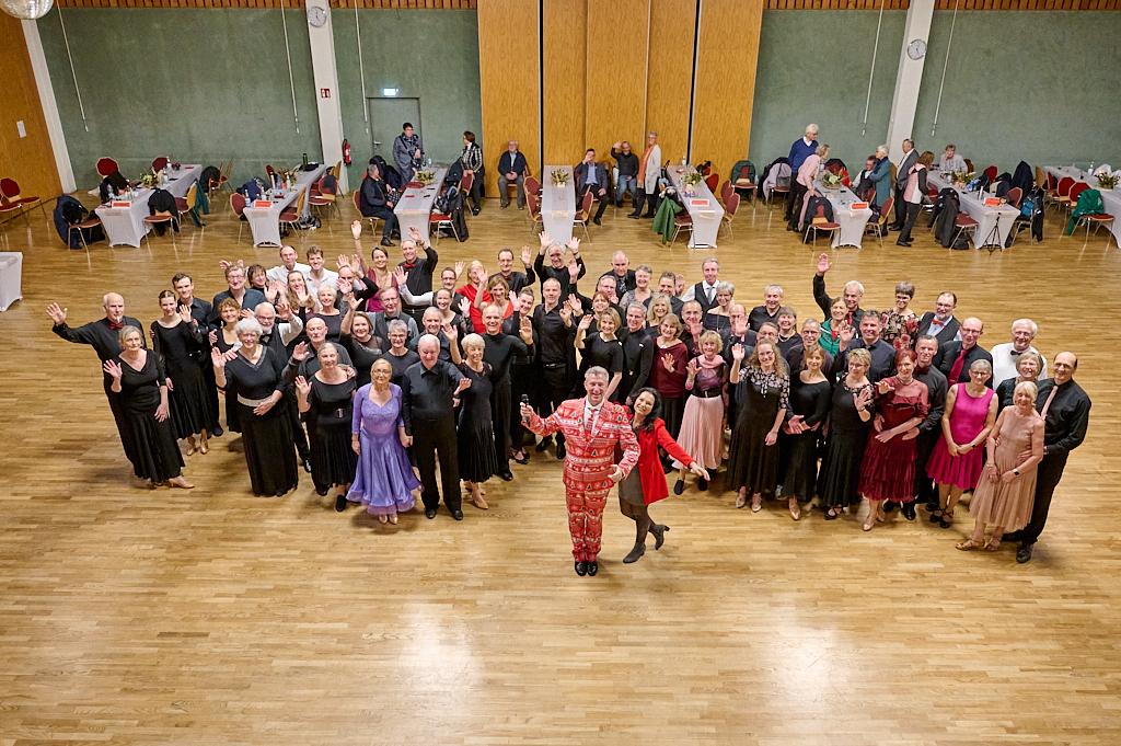 Gruppenszene von 50 tanzenden Personen in festlicher Kleidung, die gemeinsam in einem Saal posieren und l&auml;cheln.