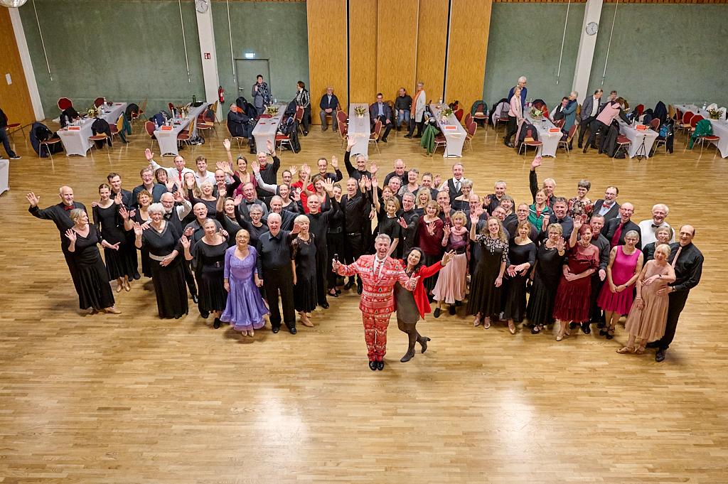 Gruppenshot von &auml;lteren T&auml;nzern in eleganter Kleidung, die fr&ouml;hlich in einem Saal mit Tischen stehen.