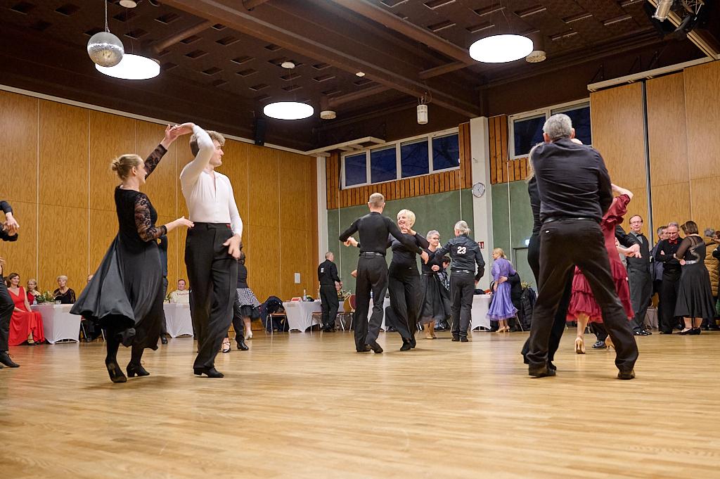 Tanzende Paare in eleganter Kleidung auf einer Holzfl&auml;che, umgeben von Tischen in einem festlichen Saal.