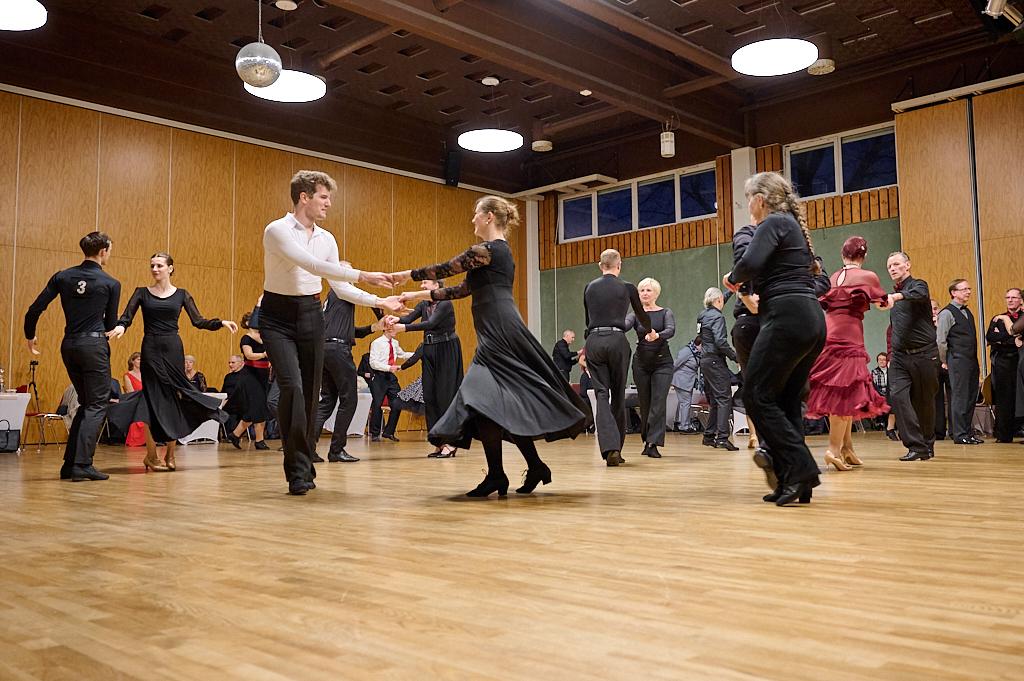 Tanzende Paare in schwarz und rot auf einer Holztanzfl&auml;che, umgeben von Zuschauern in einem festlichen Saal.