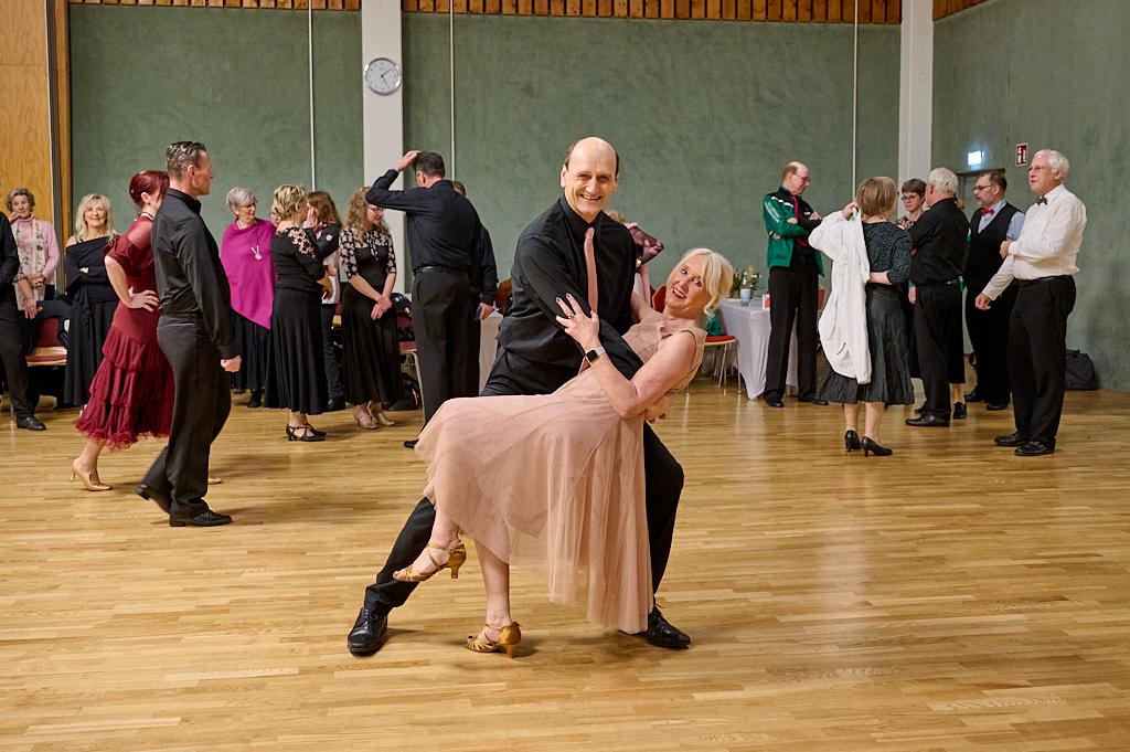 Tanzendes Paar in eleganter Kleidung, umgeben von G&auml;sten auf einer Tanzfl&auml;che in einem festlichen Saal.