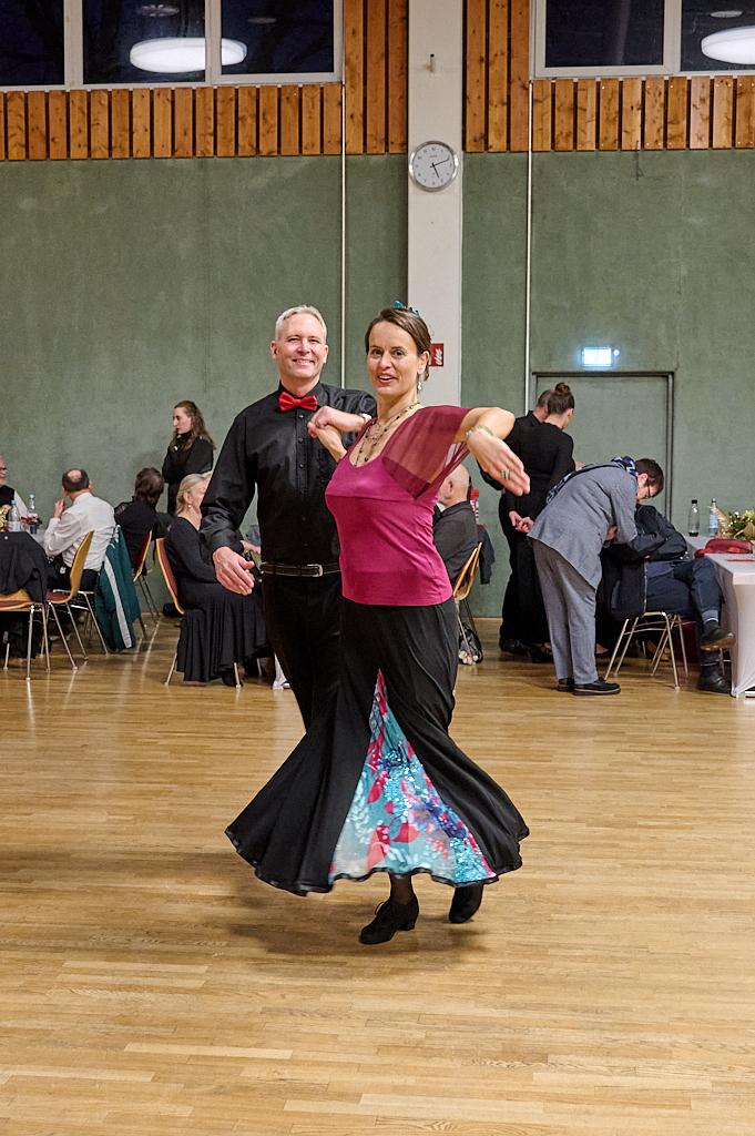 Tanzendes Paar in eleganter Kleidung, umgeben von G&auml;sten in einem festlichen Saal mit Holzw&auml;nden.