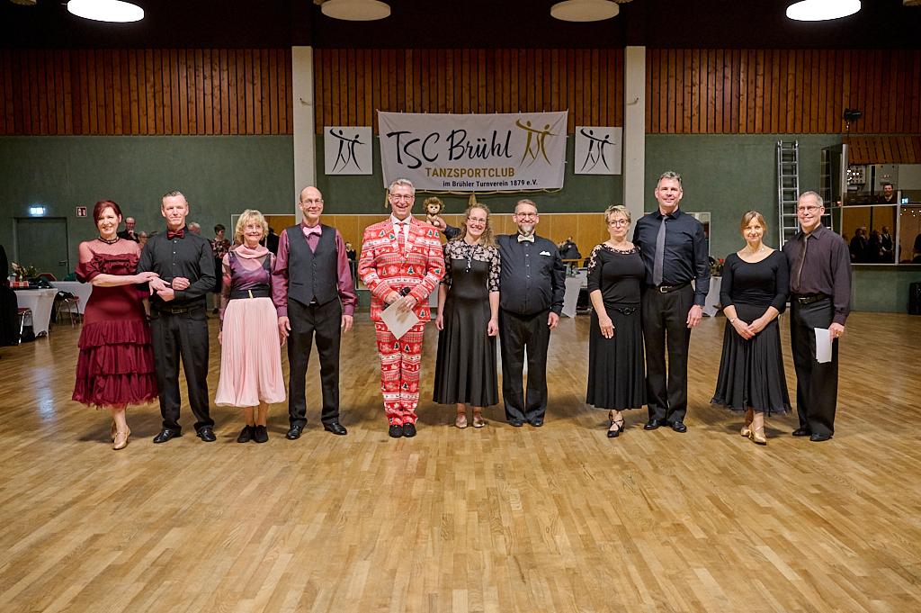 Gruppenfoto von T&auml;nzern in formeller Kleidung vor einer Wand mit dem Logo des TSC Br&uuml;hl in einem Tanzsaal.