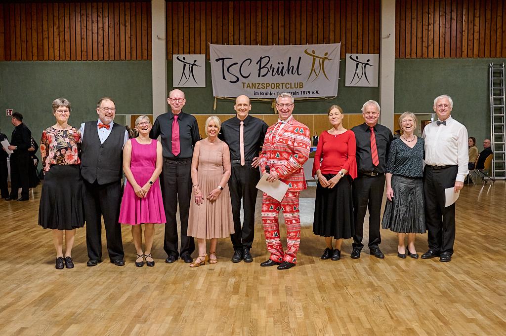 Gruppe von Tanzsportlern in formeller Kleidung, posierend vor einem Banner des TSC Br&uuml;hl im Hintergrund.