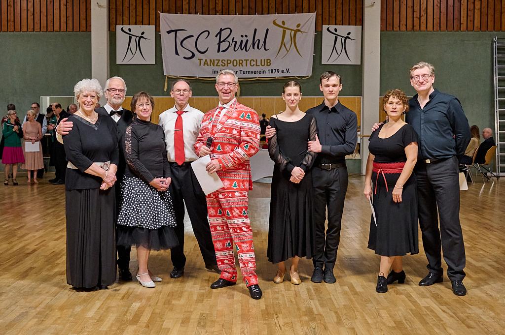 Gruppe von T&auml;nzern in festlicher Kleidung steht vor einem Tanzsaal mit TSC Br&uuml;hl Banner im Hintergrund.