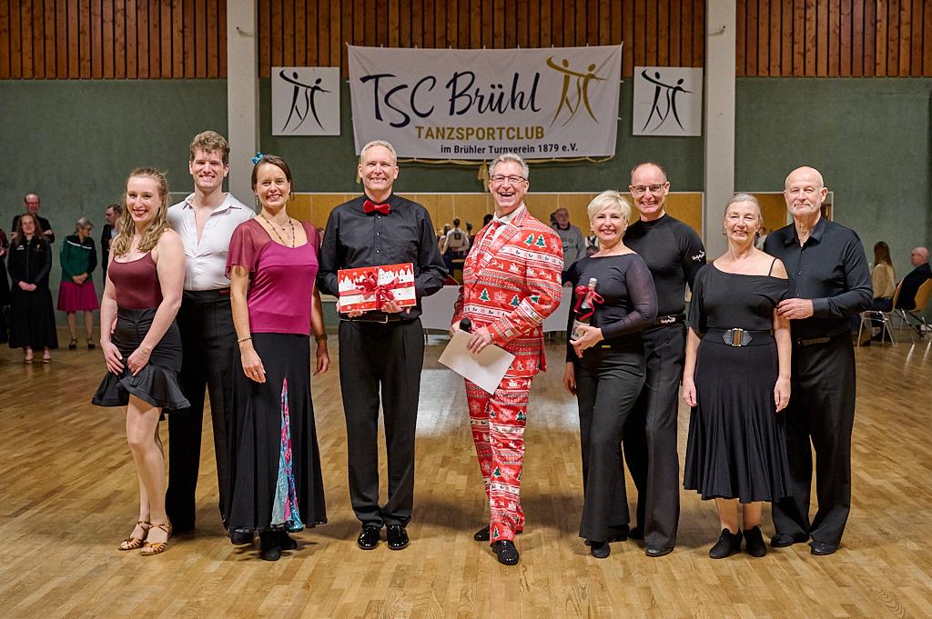 Gruppe von T&auml;nzern in festlicher Kleidung posiert l&auml;chelnd in einem Tanzsaal mit einem Banner des TSC Br&uuml;hl im Hintergrund.
