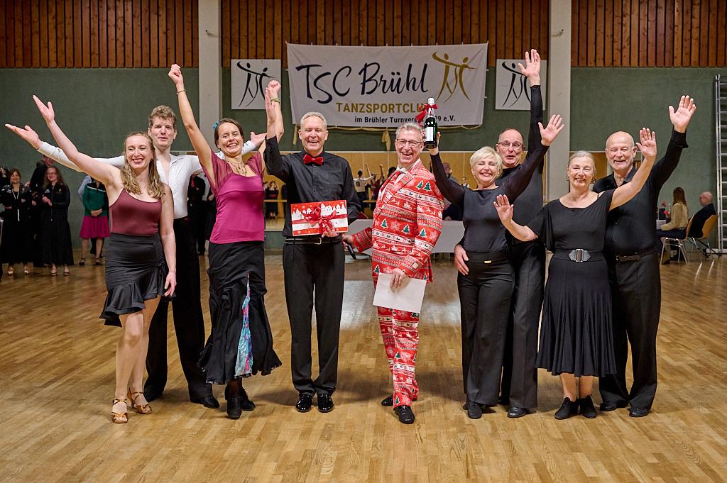 Gruppe von T&auml;nzern in festlicher Kleidung feiert mit einem Pokal in einer Tanzhalle. Banner des TSC Br&uuml;hl im Hintergrund.