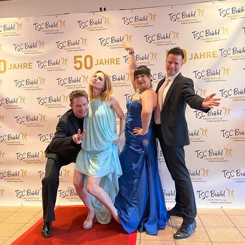 Vier Tanzsportler posieren vor einem roten Teppich und einer Feier-Wand zum 50-jährigen Jubiläum des TSC Brühl.
