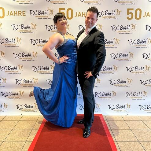 Ein Paar posiert vor einem festlichen Hintergrund mit dem Schriftzug „50 Jahre TSC Brühl“ auf einem roten Teppich.