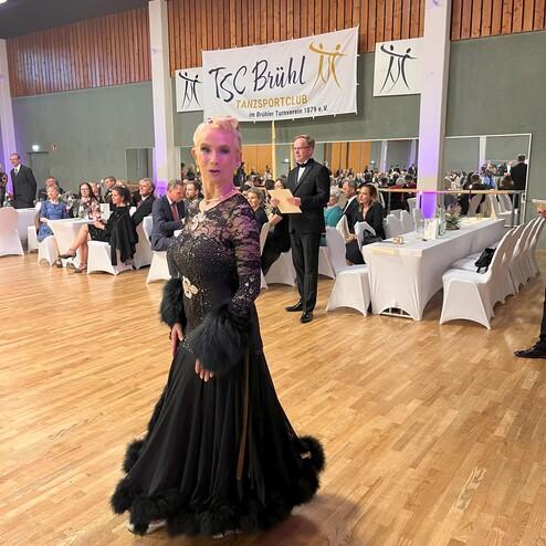 Eine Tänzerin in einem eleganten, schwarzen Kleid mit Pailletten und Pelzverzierungen steht in einem festlichen Saal.